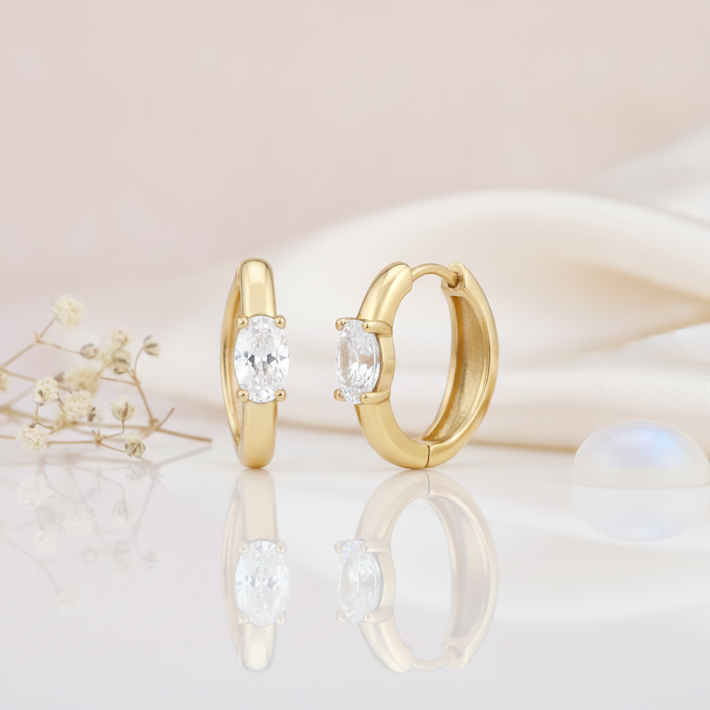 gold huggie hoop earrings with a single oval-cut White CZ stone on a pink background with dried baby's breath and a moonstone cabochon