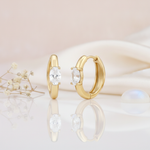 gold huggie hoop earrings with a single oval-cut White CZ stone on a pink background with dried baby's breath and a moonstone cabochon
