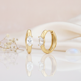 gold huggie hoop earrings with a single oval-cut White CZ stone on a pink background with dried baby's breath and a moonstone cabochon
