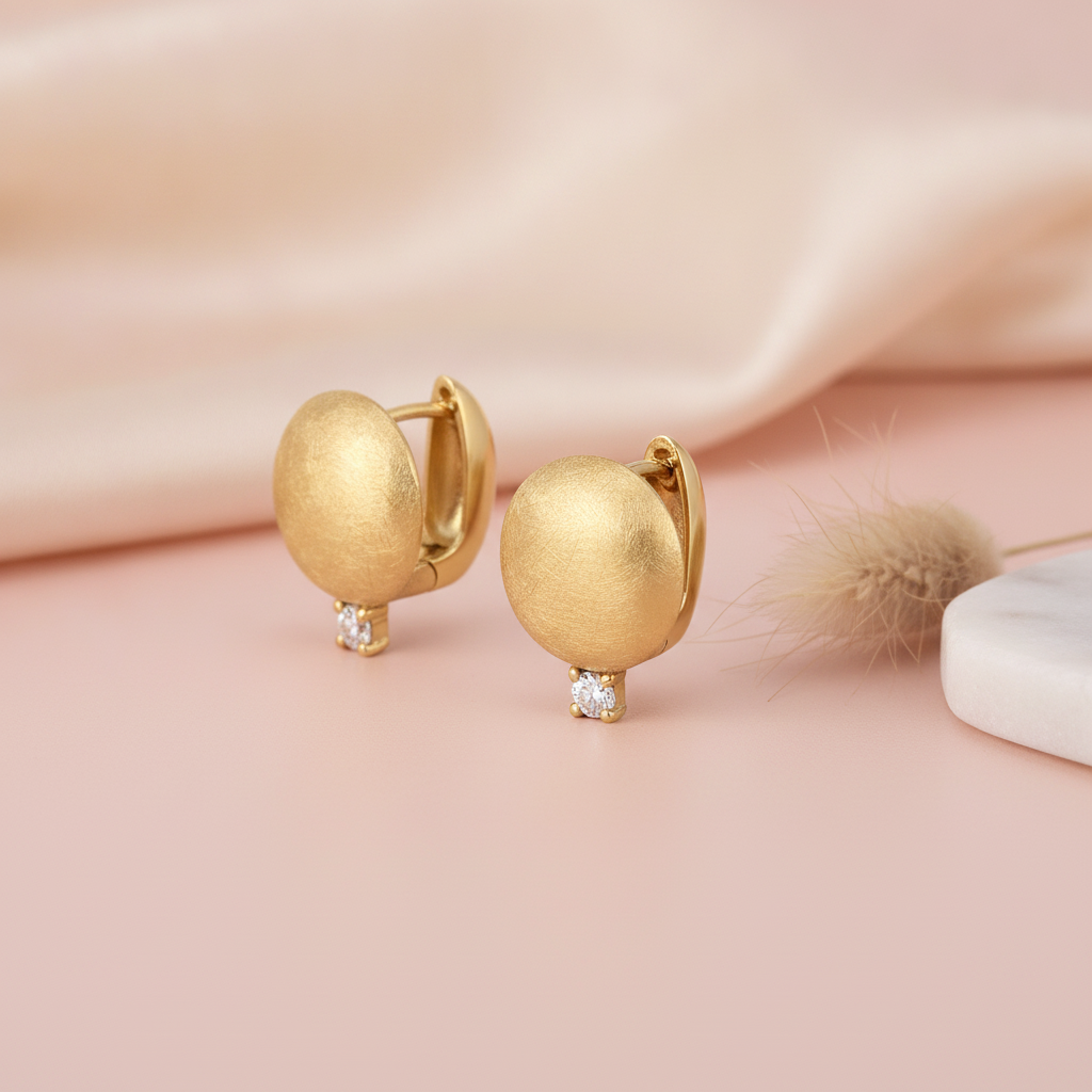 brushed gold huggie hoop earrings with a single clear stone drop, styled on a blush pink background with draped silk and a dried grass sprig