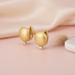 brushed gold huggie hoop earrings with a single clear stone drop, styled on a blush pink background with draped silk and a dried grass sprig
