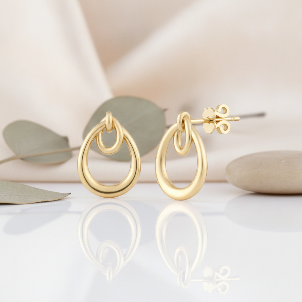 interlocking double-teardrop gold stud earrings styled on a cream background with draped silk, eucalyptus leaves, and smooth stones