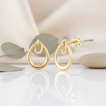 interlocking double-teardrop gold stud earrings styled on a cream background with draped silk, eucalyptus leaves, and smooth stones