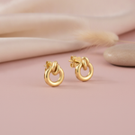 interlocking open circle gold stud earrings with a sculpted top arch, styled on a blush pink background with draped silk and a dried grass sprig