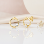 open gold ring stud earrings with a polished bead and a single clear stone, styled on a blush background with draped silk and dried flowers