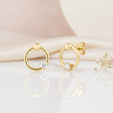open gold ring stud earrings with a polished bead and a single clear stone, styled on a blush background with draped silk and dried flowers