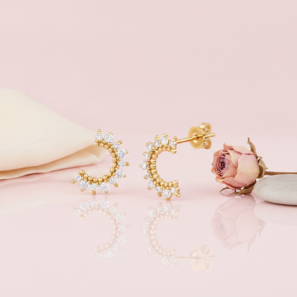 gold crescent stud earrings lined with clear stones, styled on a pink background with draped silk and a dried rose