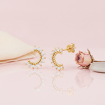 gold crescent stud earrings lined with clear stones, styled on a pink background with draped silk and a dried rose