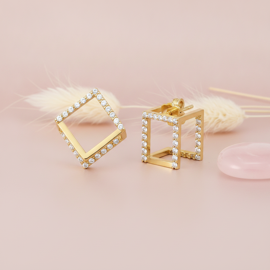 open cubic gold stud earrings lined with clear stones, styled on a pink background with a dried grass sprig and a pink stone