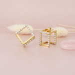 open cubic gold stud earrings lined with clear stones, styled on a pink background with a dried grass sprig and a pink stone