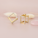 open cubic gold stud earrings lined with clear stones, styled on a pink background with a dried grass sprig and a pink stone