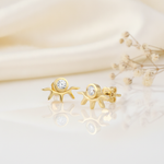 gold sunburst stud earrings with a bezel-set clear stone above radiating rays, styled on an ivory background with draped silk and dried flowers