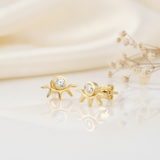 gold sunburst stud earrings with a bezel-set clear stone above radiating rays, styled on an ivory background with draped silk and dried flowers
