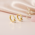 gold huggie hoop earrings with two cascading clear stones, styled on a blush background with draped silk and a pink stone