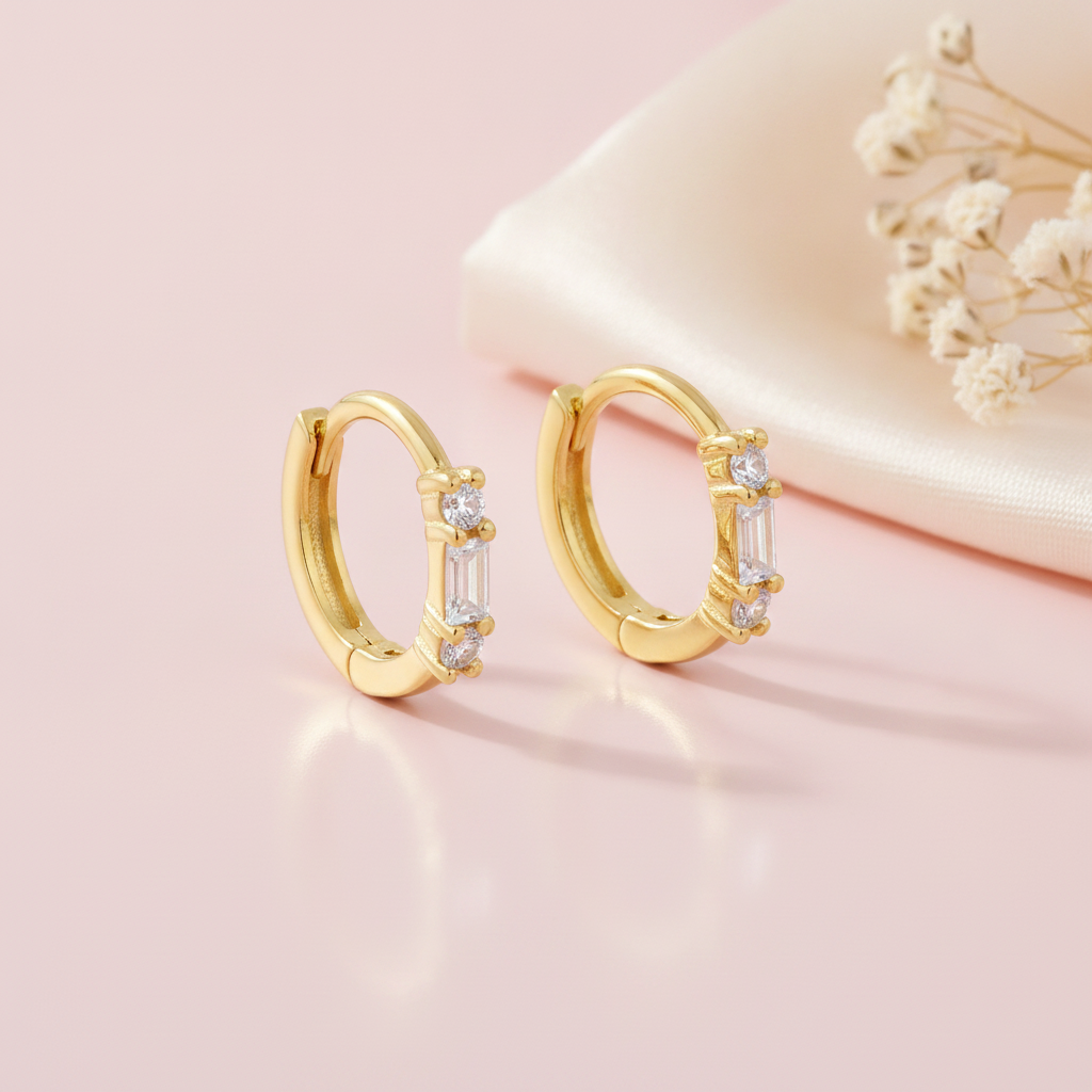 gold huggie hoop earrings with a baguette-cut stone flanked by round clear stones, styled on a pink background with draped silk