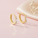 gold huggie hoop earrings with a baguette-cut stone flanked by round clear stones, styled on a pink background with draped silk