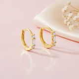 gold huggie hoop earrings with a baguette-cut stone flanked by round clear stones, styled on a pink background with draped silk