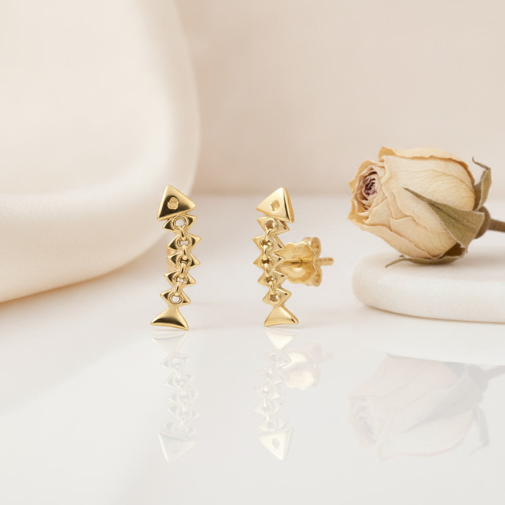 articulated gold fishbone stud earrings with linked chevron segments, styled on a cream background with draped silk and a dried rose