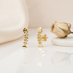 articulated gold fishbone stud earrings with linked chevron segments, styled on a cream background with draped silk and a dried rose