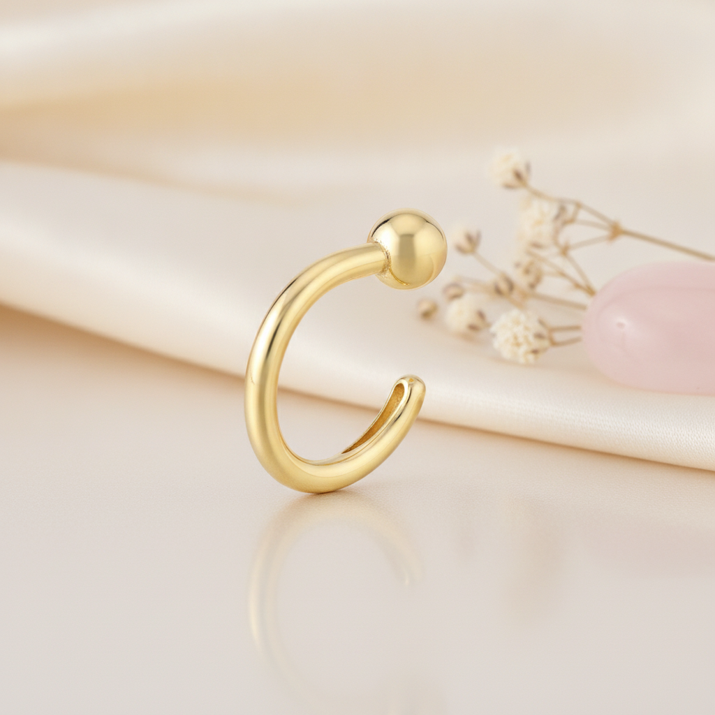 open gold arc stud earring with a single sphere end, styled on a cream background with draped silk and a pink stone