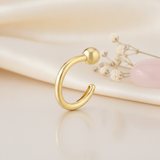 open gold arc stud earring with a single sphere end, styled on a cream background with draped silk and a pink stone