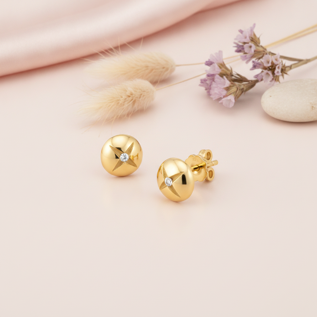 gold dome stud earrings etched with a star and set with a single clear stone, styled on a pink background with dried grass and purple statice flowers