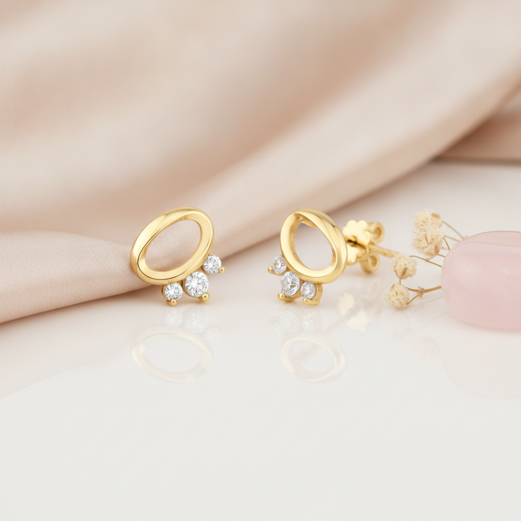 open oval gold stud earrings with three clear stones at the base, styled on a cream background with draped silk and a pink stone