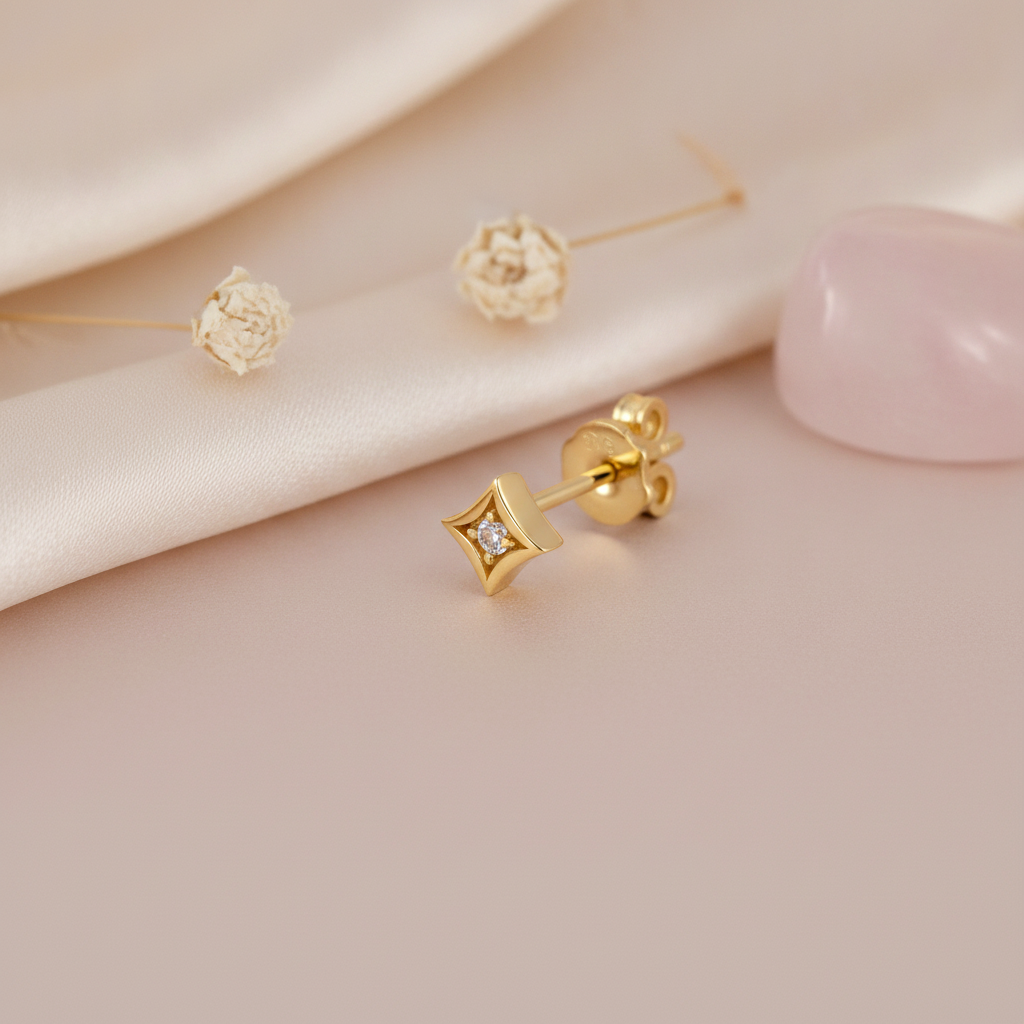 small gold star-shaped stud earring with a single clear stone, styled on a pink background with dried flower puffs and a pink stone