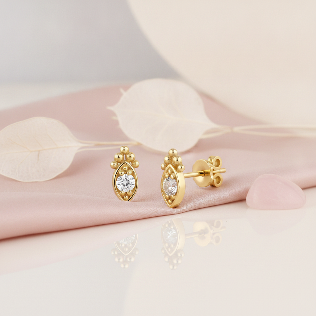 almond-shaped gold stud earring with a central clear stone and beaded crest, styled on a pink background with dried silvery leaves and a pink stone