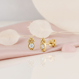 almond-shaped gold stud earring with a central clear stone and beaded crest, styled on a pink background with dried silvery leaves and a pink stone