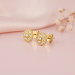 circular gold medallion stud earrings with a star center and clear stones, styled on a pink background with dried baby's breath flowers