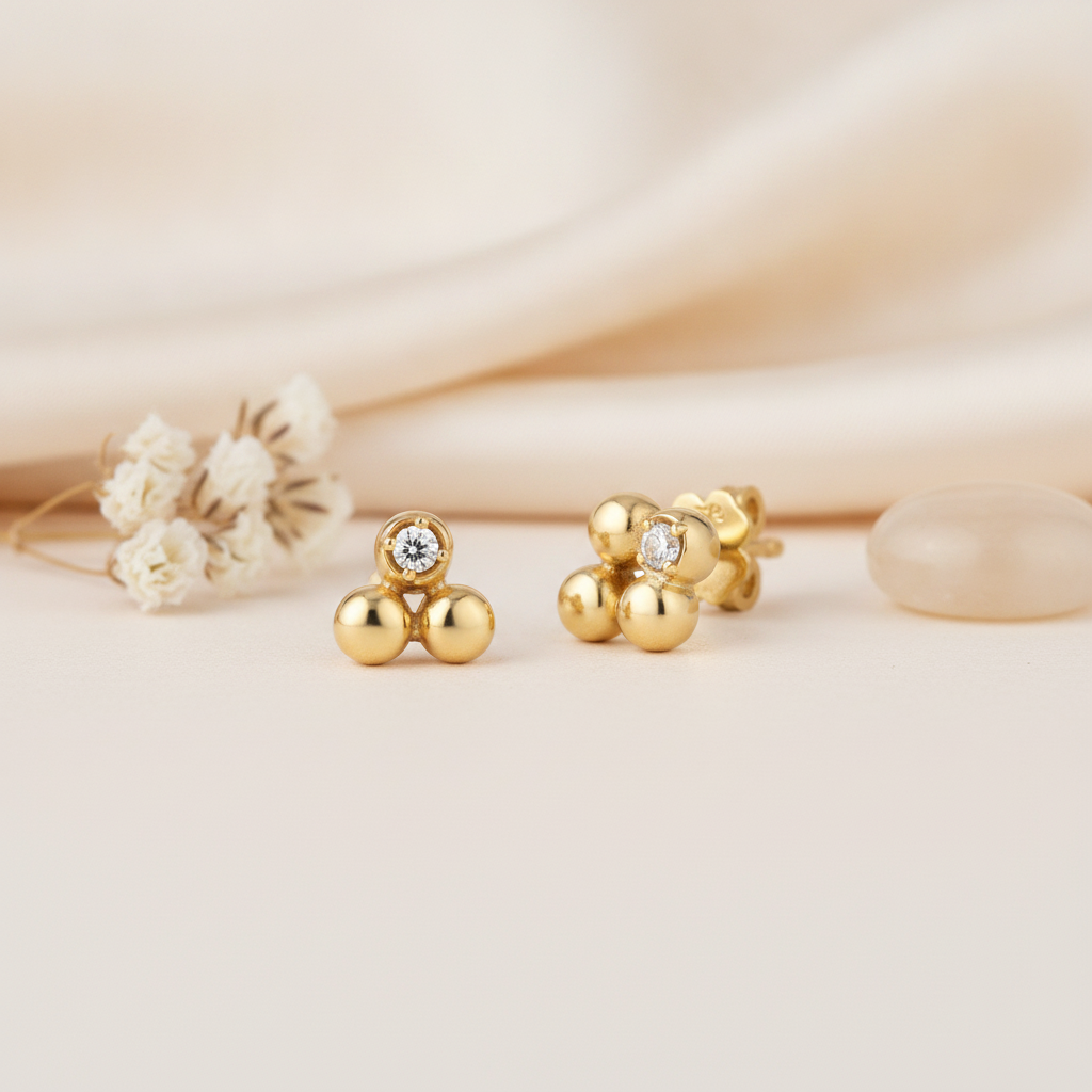 trio of polished gold sphere stud earrings with a single clear stone accent, styled on a cream background with dried flowers and a translucent stone