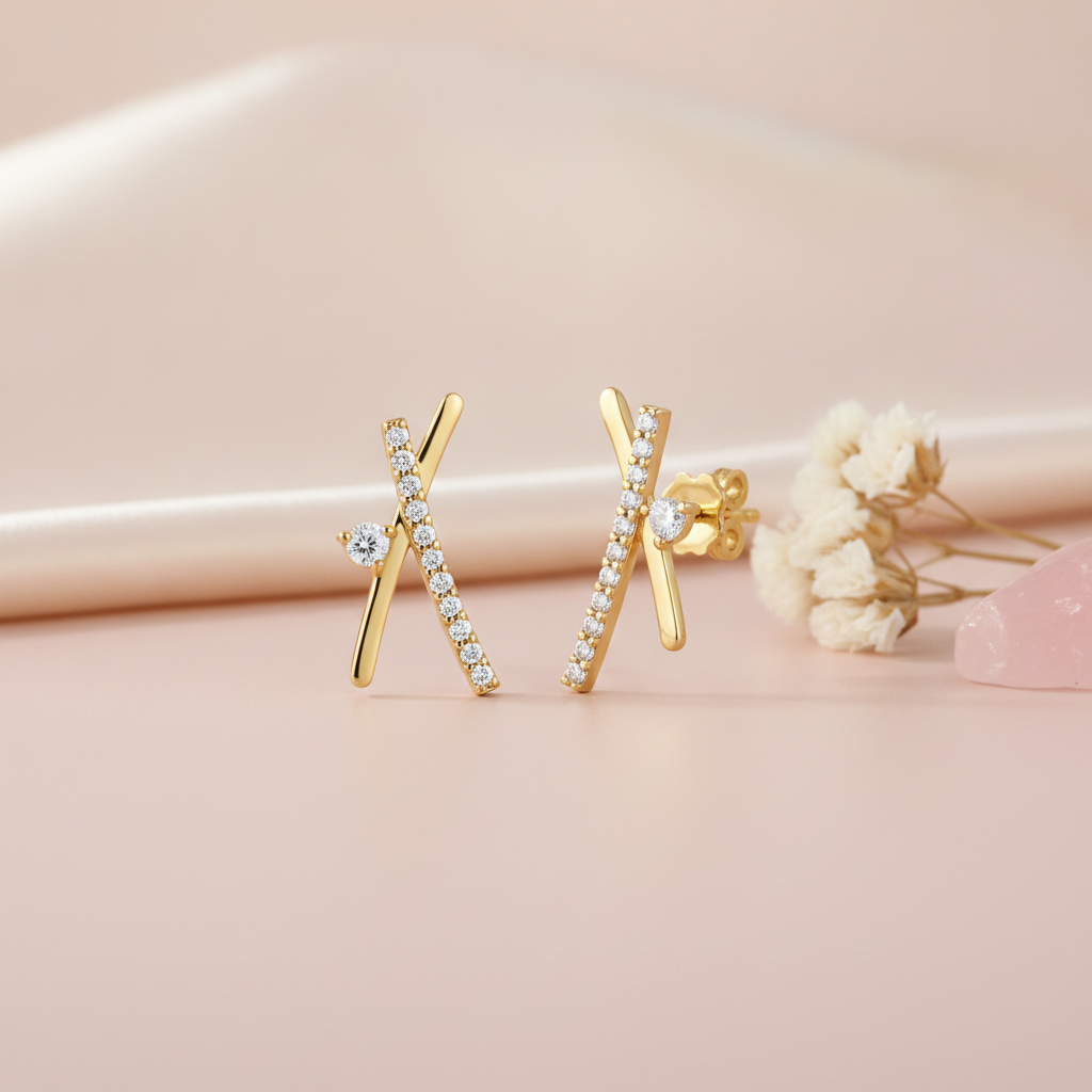 crossed gold bar stud earrings with pave clear stones and a single round stone accent, styled on a pink background with dried flowers and a pink stone