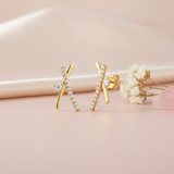 crossed gold bar stud earrings with pave clear stones and a single round stone accent, styled on a pink background with dried flowers and a pink stone