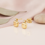 soft-edged gold cushion stud earrings with a single clear stone above, styled on a pink background with draped silk and eucalyptus leaves
