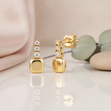 gold stud earrings with a vertical trio of clear stones above a smooth cushion drop, styled on a pink background with eucalyptus leaves and a stone