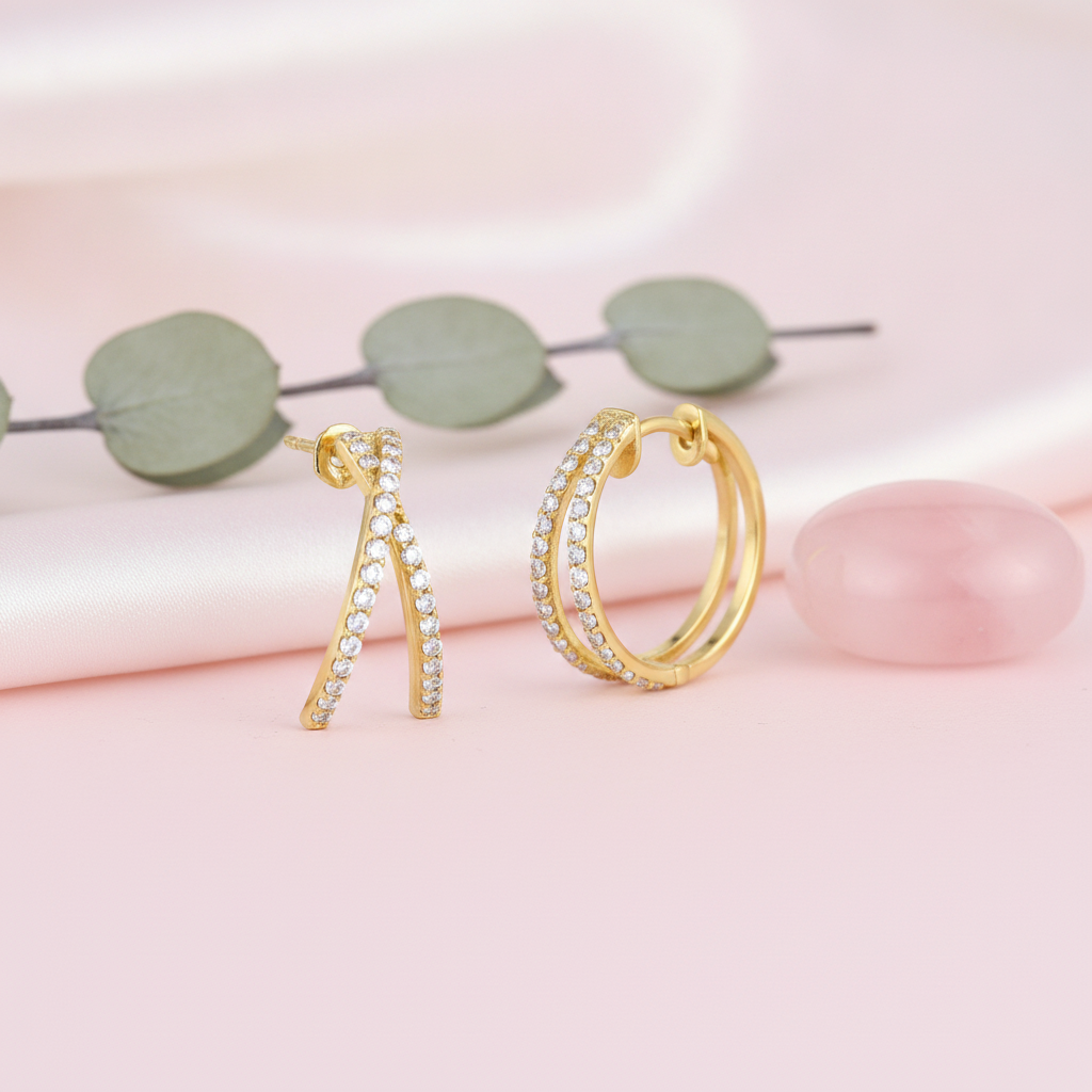 criss-crossing gold hoop earrings set with clear stones, styled on a pink background with eucalyptus leaves and a pink stone