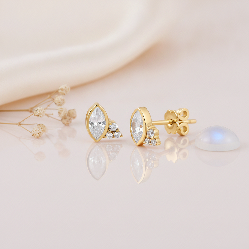 gold stud earrings with a marquise-cut clear stone and a trio of small clear stones, styled on a cream background with dried flowers and a moonstone cabochon