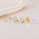 gold stud earrings with a marquise-cut clear stone and a trio of small clear stones, styled on a cream background with dried flowers and a moonstone cabochon