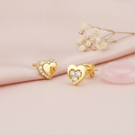 gold heart stud earrings with a polished center and an arc of clear stones, styled on a pink background with dried flowers and a pink stone