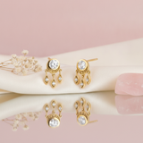 gold stud earrings with a beaded halo center and three dangling leaf charms set with clear stones, styled on a pink and cream background with dried flowers and a pink stone