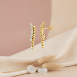 gold earrings with a curved row of clear stones beside a strand of polished gold beads, styled with dried flowers and moonstone beads on a blush background