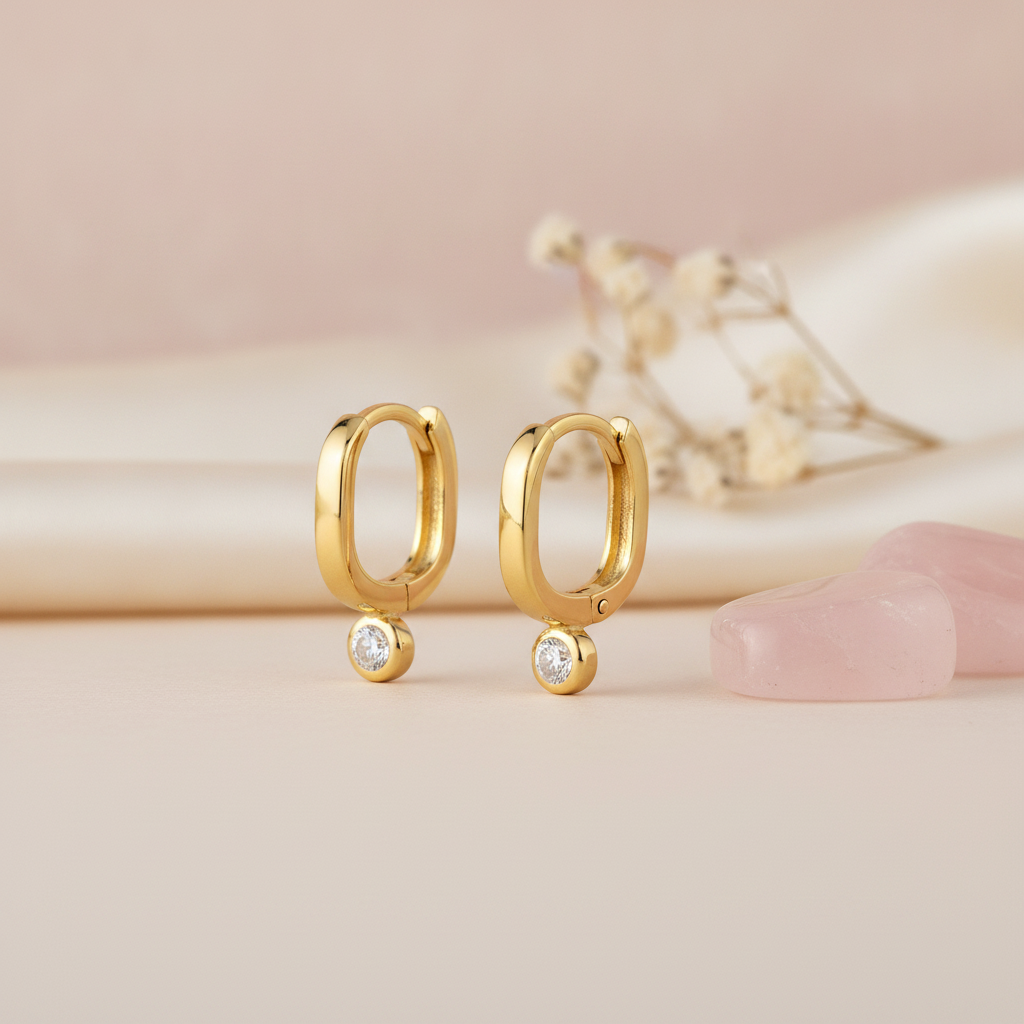 gold oval huggie earrings with a single bezel-set clear stone, styled with dried baby's breath and pink stones on a blush background