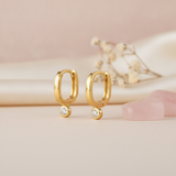 gold oval huggie earrings with a single bezel-set clear stone, styled with dried baby's breath and pink stones on a blush background