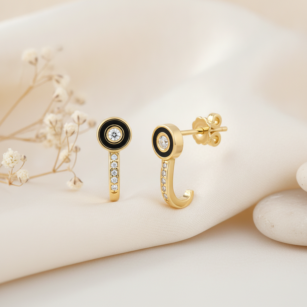 gold J-hook earrings with a black enamel halo around a center stone and a pave curved drop, styled with dried baby's breath and stone pebbles on a cream background