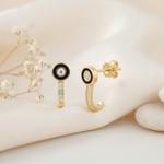 gold J-hook earrings with a black enamel halo around a center stone and a pave curved drop, styled with dried baby's breath and stone pebbles on a cream background
