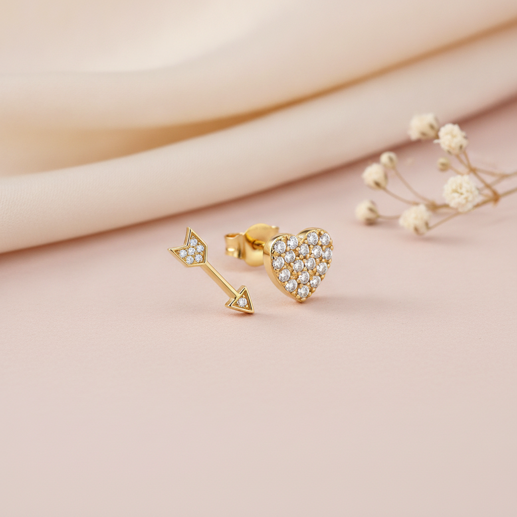 gold asymmetric stud earrings featuring a pave heart and a pave arrow, styled with dried baby's breath flowers on a blush background