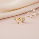gold asymmetric stud earrings featuring a pave heart and a pave arrow, styled with dried baby's breath flowers on a blush background