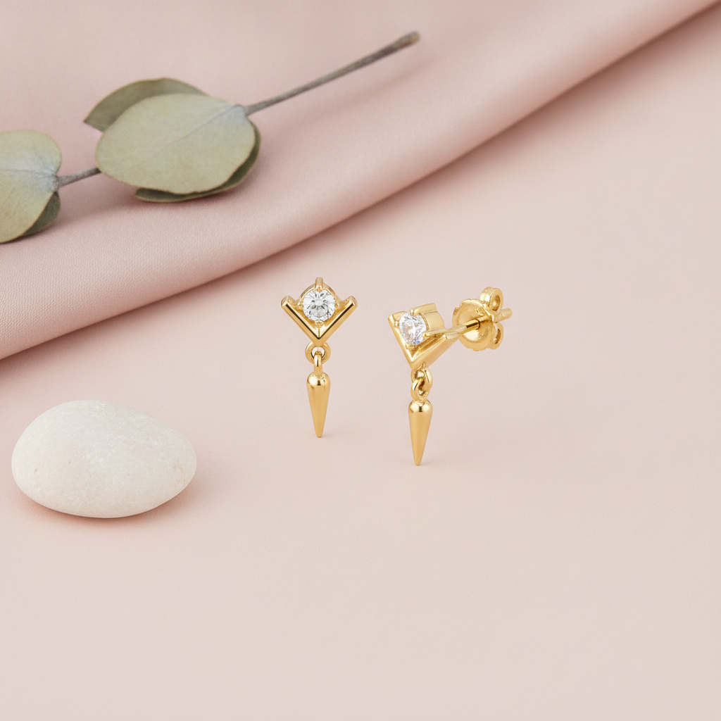 gold chevron stud earring with a clear stone and dangling spike drop, styled with a smooth white stone on a pink background