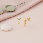 gold chevron stud earring with a clear stone and dangling spike drop, styled with a smooth white stone on a pink background